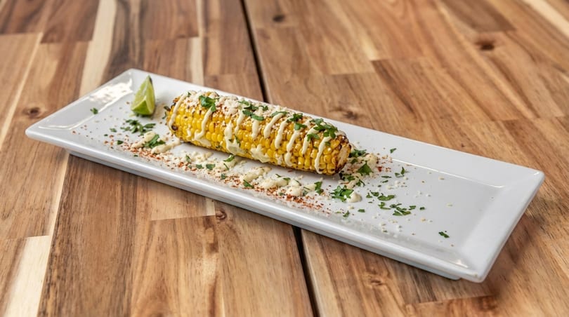 Elote, Mexican street corn, on a white rectangular plate with a lime wedge on a wooden table.