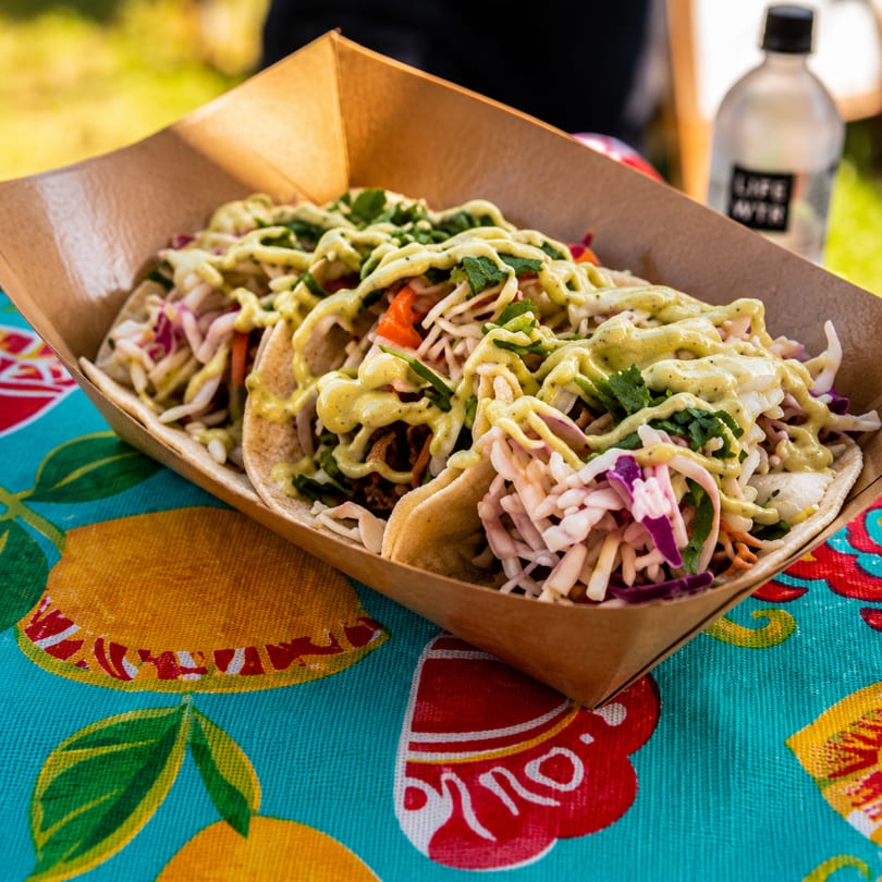Three tacos with green sauce and slaw in a cardboard tray on a colorful tablecloth.