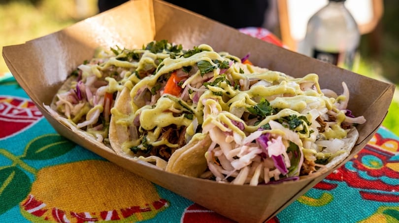 Three tacos with meat, cabbage, cilantro, and a creamy green sauce in a cardboard tray.
