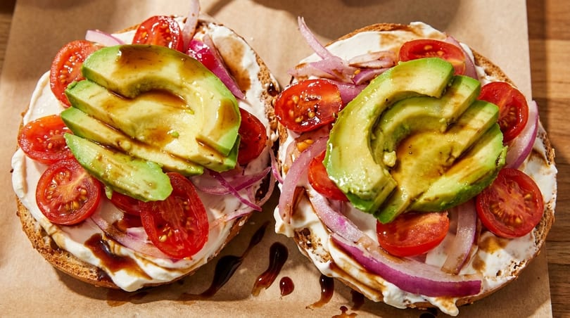 Two bagels topped with cream cheese, sliced avocado, cherry tomatoes, red onion, and balsamic glaze.