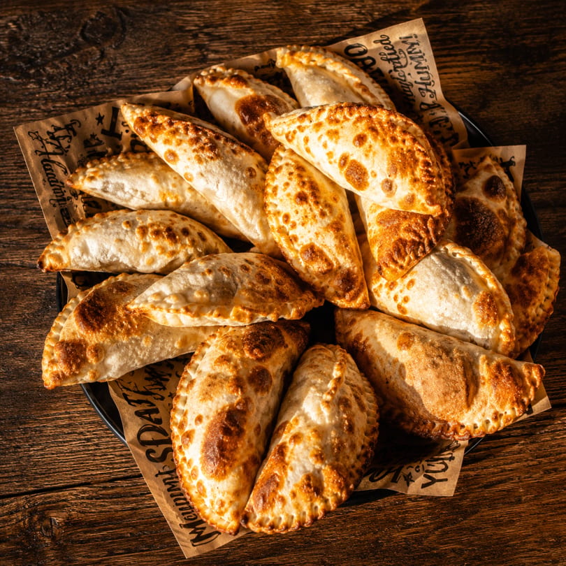A platter of golden-brown empanadas, some with crimped edges, on branded parchment paper.