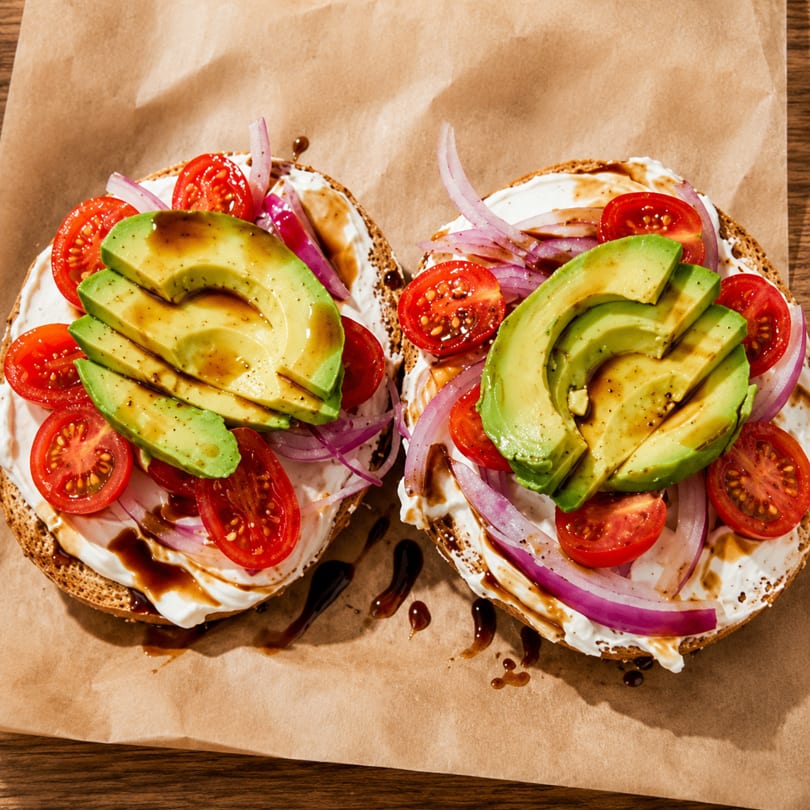 Two bagels topped with cream cheese, avocado, cherry tomatoes, red onion, and balsamic glaze.