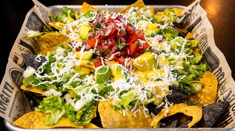 A tray of colorful nachos with cheese, lettuce, avocado, pico de gallo, and crumbled white cheese.