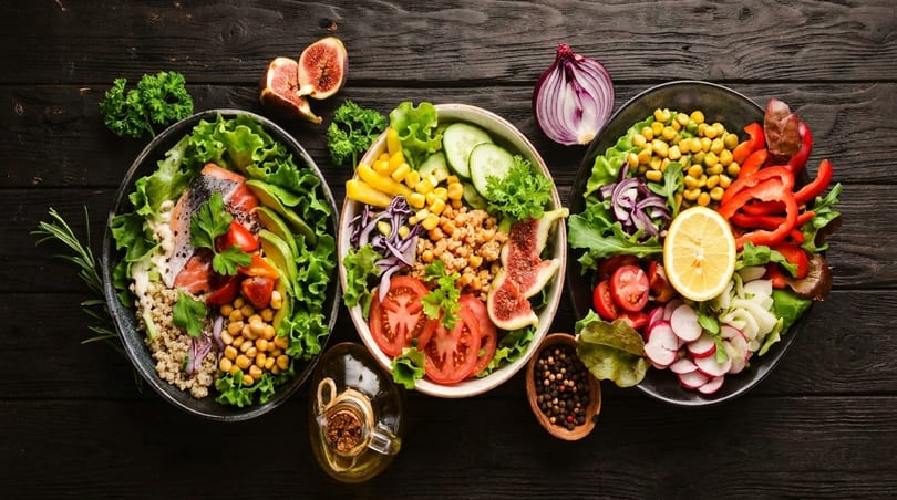 Three colorful, healthy salad bowls with various fresh ingredients on a dark wooden table.
